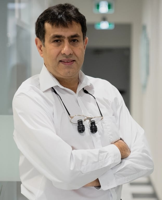 Dr. Ali Mandoub Hashemi, qualified dentist at Hastings Dental Center, wearing dental loupes, standing confidently in a modern dental office setting.